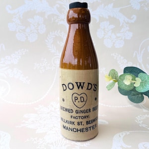 May include: A brown and cream ceramic bottle with a black cap. The label reads DOWD'S P.D. BREWED GINGER BEER FACTORY: MILLKIRK ST, BESWICK MANCHESTER. The bottle has a cylindrical body and a narrow neck, featuring a glossy finish on the upper portion.