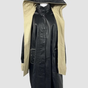 Puede incluir: Un abrigo con capucha de cuero negro con interior beige. El abrigo tiene cierre de botones, mangas largas y dos bolsillos delanteros. La capucha y el interior son de color beige claro, creando un contraste con el exterior negro.