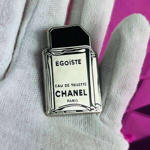 May include: A silver-colored perfume bottle-shaped pin with the text "&Eacute;GO&Iuml;STE" and "CHANEL PARIS" in black. The pin has a black cap and is set against a white background.