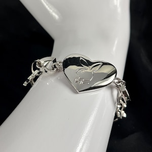 Puede incluir: Una pulsera plateada con un dije en forma de corazón con un logotipo de conejo. La pulsera tiene un diseño de eslabones de cadena y un cierre de broche. El dije de corazón es el punto focal, con el logotipo del conejo grabado.