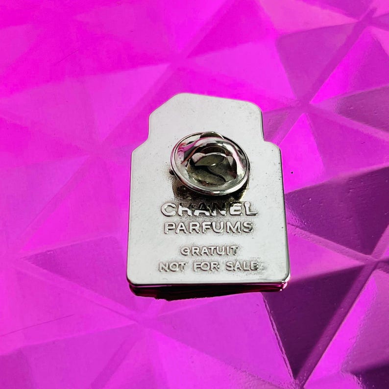 May include: A silver-colored pin in the shape of a perfume bottle. The pin has the words "CHANEL PARFUMS GRATUIT NOT FOR SALE" engraved on it. The pin is set against a bright pink, textured background.