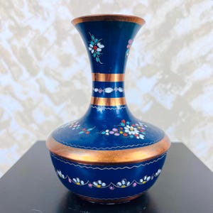 Könnte beinhalten: Eine tiefblaue Metallvase mit einem ausgestellten Hals und einem abgerundeten Körper. Die Oberfläche ist mit handgemalten Blumenmotiven in Weiß, Rosa und Hellblau verziert, akzentuiert durch horizontale kupferfarbene Bänder am Hals und Fuß.