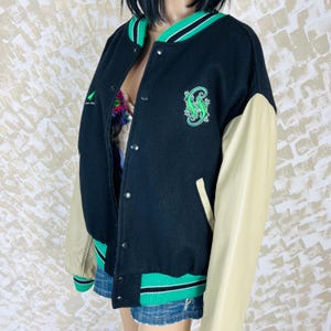 May include: A black varsity jacket with beige sleeves, featuring a green lizard embroidery on the chest. The jacket has a snap-button front closure, a ribbed collar, and ribbed cuffs with green and white stripes. It is worn over a patterned top.