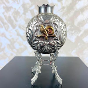 May include: A silver-toned spherical vessel with a crown-shaped rim, featuring an embossed leaf pattern and a central gold-toned bird perched on a branch. The vessel is supported by a decorative silver-toned metal stand with three ornate legs.