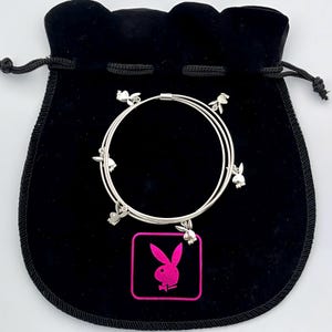 Puede incluir: Un brazalete plateado con varios abalorios en forma de conejo. El brazalete está en una bolsa de terciopelo negro con un logotipo de conejo Playboy rosa.