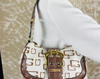May include: A white and brown shoulder bag featuring a repeating G logo pattern, a prominent central brown strap with a large gold-tone buckle, gold-tone metal studs along the edges, and a single shoulder strap with gold-tone hardware.