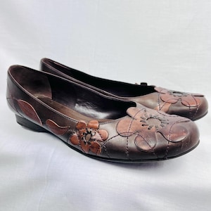 May include: Brown leather flats featuring floral cut-out detailing. The shoes have a low heel and decorative stitching. The flowers are a darker shade of brown, providing contrast. The shoes are presented on a white backdrop.