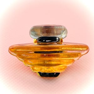 Op de afbeelding: Een decoratieve glazen parfumfles met een gelaagde, amberkleurige body en een doorzichtige, ovaalvormige stopper. De basis is zwart en de fles heeft een uniek, artistiek ontwerp. De achtergrond is zachtroze.