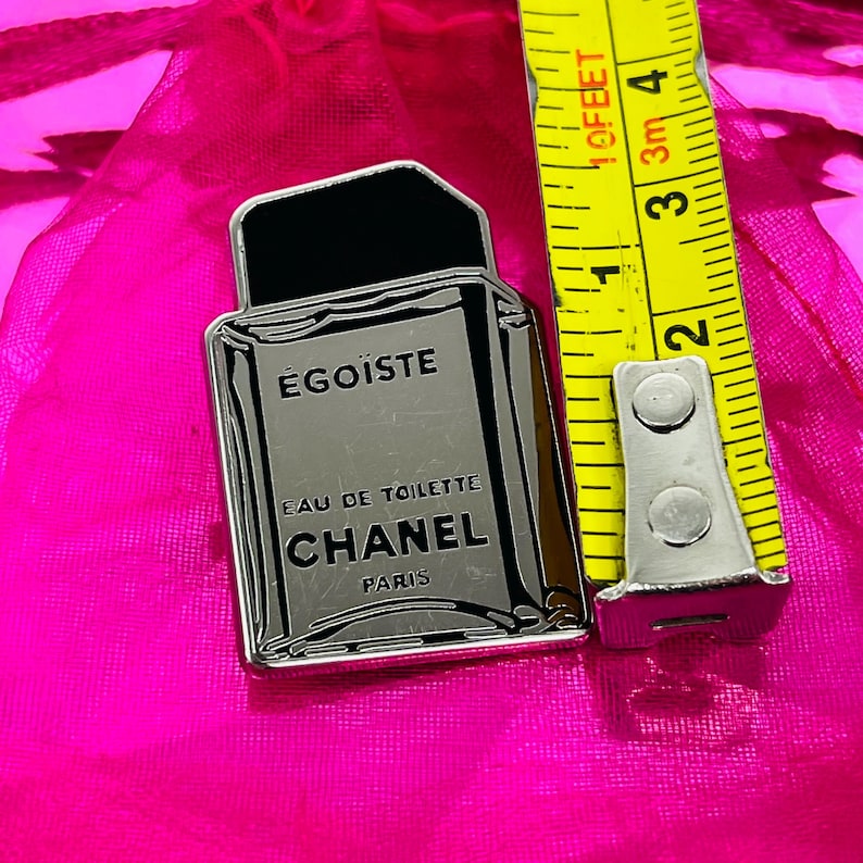 May include: A silver-colored perfume bottle-shaped pin with the word "&Eacute;GO&Iuml;STE" at the top, followed by "EAU DE TOILETTE" and "CHANEL PARIS". The pin is next to a yellow measuring tape, and the background is pink.