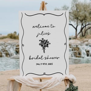 May include: A white sign with black text and a floral illustration on an easel. The sign reads "Welcome to Julie's Bridal Shower July 9th, 2025".