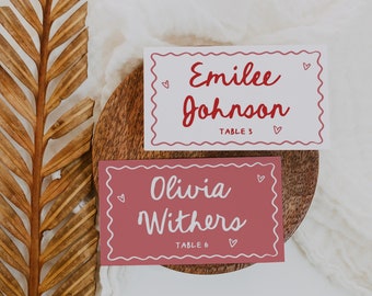 May include: Two place cards with red script names and table numbers on a wooden surface. The top card says "Emilee Johnson, Table 3" and the bottom card says "Olivia Withers, Table 6".