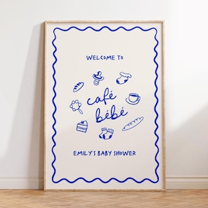 May include: A white poster with a blue border and blue text that says "Welcome to cafe bebe Emily's Baby Shower". The poster has a blue illustration of a pacifier, a pair of socks, a cup of coffee, a loaf of bread, a cake, a rattle, and a pair of socks.