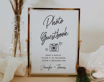 May include: A gold-framed sign with white background and black text that reads "Photo Guestbook" with a camera icon and hearts. The text below reads "Snap a photo, place it in the book, leave a message for Jennifer + Thomas".
