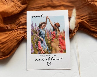 May include: A white card with the text "sarah, will you be my maid of honor?" and a heart symbol. The card has a photo of two people in a field of flowers.