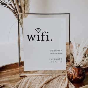 May include: A framed white sign with the word "wifi" in a bold, black font, and a wifi symbol. Below, it says "NETWORK" and "PASSWORD" with spaces for details. The sign is in a gold frame, with decorative dried plants in the background.