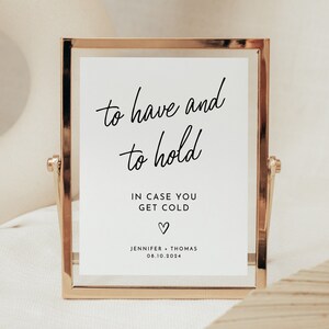 May include: A gold-framed sign with the text "to have and to hold" in black cursive script. Below the text is "IN CASE YOU GET COLD" and a small heart symbol. The bottom of the sign reads "JENNIFER + THOMAS 08.10.2024".