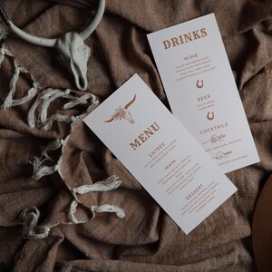 Puede incluir: Una tarjeta blanca con la palabra "MENU" impresa en texto marrón. La tarjeta tiene una silueta marrón de una cabeza de toro con un diseño floral. La tarjeta está sobre un fondo de tela marrón con una calavera blanca y un sombrero marrón.