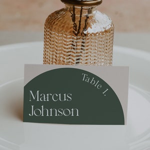 May include: A white place card with a green semi-circle design. The text "Table 1." is in the upper right corner of the semi-circle. The text "Marcus Johnson" is in the lower left corner of the semi-circle.