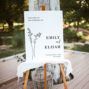May include: White wedding welcome sign with black text and a simple line drawing of flowers. The text reads "Welcome to the wedding of Emily and Elijah, Canyon Point, Utah, 04.12.2021".