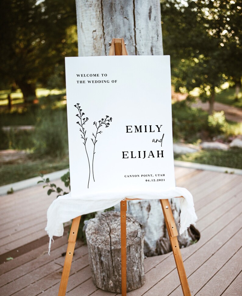May include: White wedding welcome sign with black text and a simple line drawing of flowers. The text reads "Welcome to the wedding of Emily and Elijah, Canyon Point, Utah, 04.12.2021".