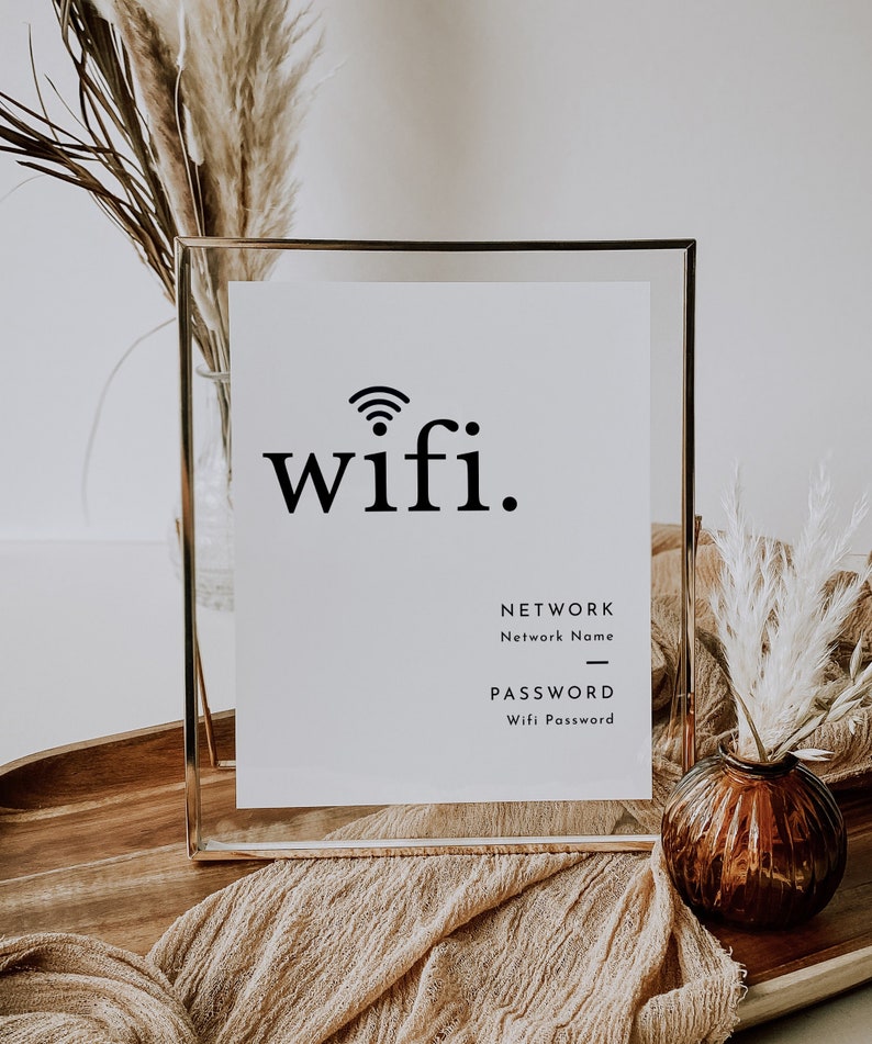 May include: A framed white sign with the word "wifi" in a bold, black font, and a wifi symbol. Below, it says "NETWORK" and "PASSWORD" with spaces for details. The sign is in a gold frame, with decorative dried plants in the background.