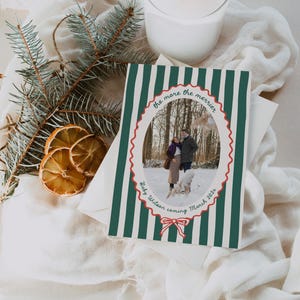 Può includere: Un biglietto a tema festivo con uno sfondo a righe verdi e bianche. Il biglietto presenta una foto di una coppia con un cane in un ambiente innevato, incorniciata da un ovale smerlato rosso. Il testo include "the more the merrier" e "Baby Wilson coming March 2024."