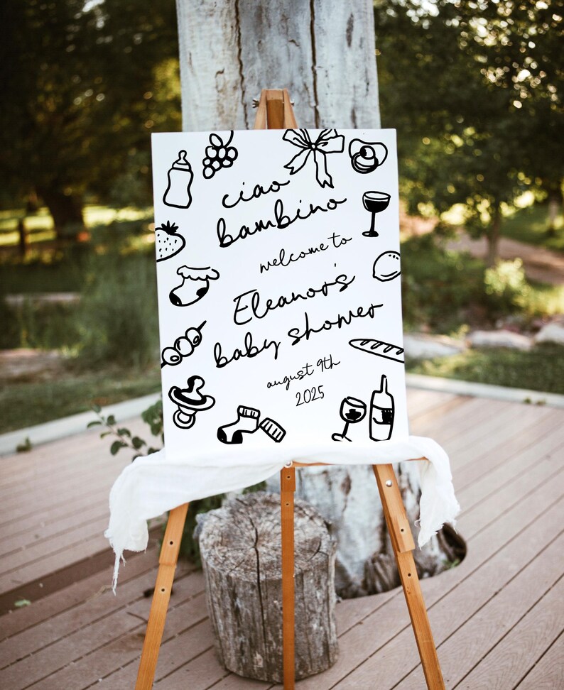 Op de afbeelding: Een wit bord met zwarte inkttekeningen van babygerelateerde items, waaronder een fles, een speen, een sok, een wijnglas en een stokbrood. De tekst op het bord luidt "Ciao bambino, welkom op Eleanor's babyshower, 9 augustus 2025".