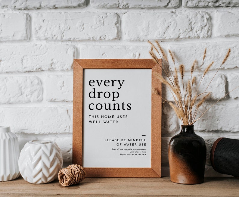 May include: A framed sign with the text "every drop counts" and "This home uses well water." The sign also includes a message about being mindful of water use. The sign is on a white brick wall with a vase of dried flowers and other decorative items.