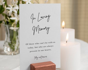 May include: A memorial card with the text "In Loving Memory" is displayed in a copper-colored wooden holder. The card includes a message about remembering loved ones. Two white candles are lit in the background, with white flowers in a vase.