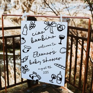 Op de afbeelding: Een wit bord met zwarte doodle-illustraties die een babyshower voor Eleanor aankondigen. Het bord zegt "Ciao bambino, welkom bij Eleanor's babyshower, 9 augustus 2025".