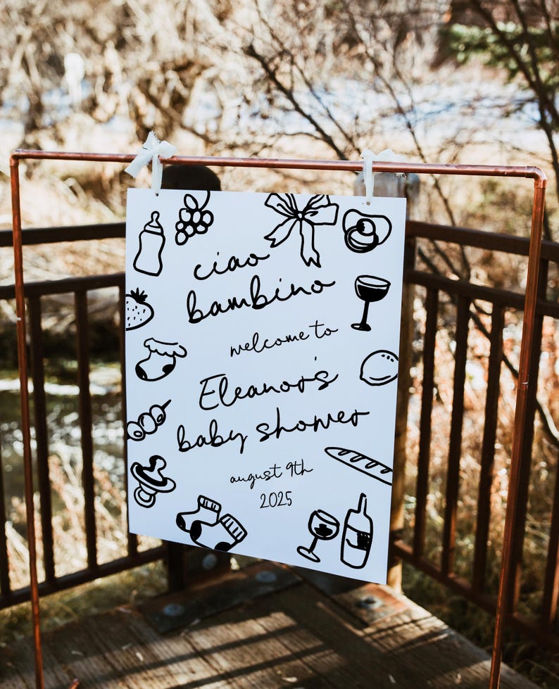 Op de afbeelding: Een wit bord met zwarte doodle-illustraties die een babyshower voor Eleanor aankondigen. Het bord zegt "Ciao bambino, welkom bij Eleanor's babyshower, 9 augustus 2025".