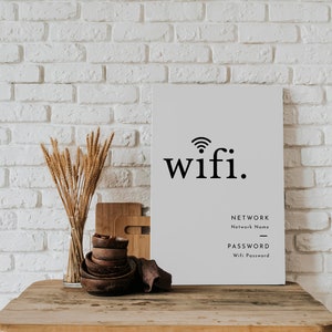 May include: A rectangular white sign with the word "wifi." in black, along with fields for network name and password. The sign is on a wooden table with decorative bowls, cutting boards, and a vase of dried wheat. The background is a white brick wall.