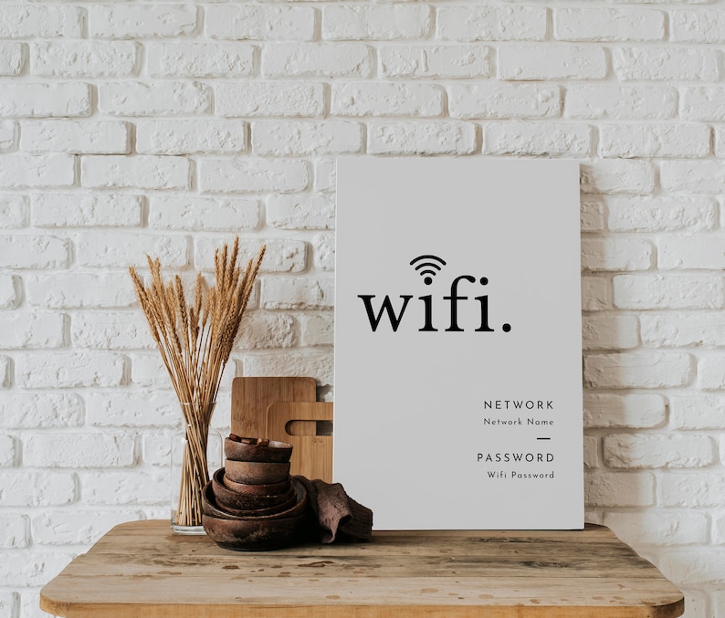 May include: A rectangular white sign with the word "wifi." in black, along with fields for network name and password. The sign is on a wooden table with decorative bowls, cutting boards, and a vase of dried wheat. The background is a white brick wall.