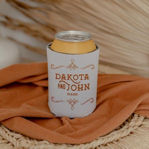 May include: White can cooler with a brown inscription that reads "Dakota and John 09.28.2023". The cooler is sitting on a brown fabric surface.