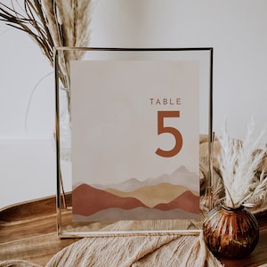 May include: A framed table number 5 with a minimalist mountain landscape design in shades of brown, beige, and gold. The frame is gold and the background is a soft beige. Decorative elements include pampas grass and a brown glass vase.