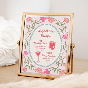Puede incluir: Un letrero con marco dorado con fondo rosa y detalles florales. El letrero muestra "Signature Drinks" con las recetas "His Whiskey Sour" y "Hers Dirty Martini". El letrero est&aacute; sobre una superficie de madera.
