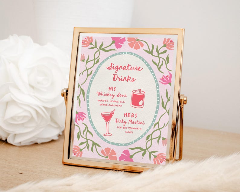 Puede incluir: Un letrero con marco dorado con fondo rosa y detalles florales. El letrero muestra "Signature Drinks" con las recetas "His Whiskey Sour" y "Hers Dirty Martini". El letrero est&aacute; sobre una superficie de madera.