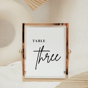 May include: Gold-framed table number sign with a white background and black text that reads "TABLE three".