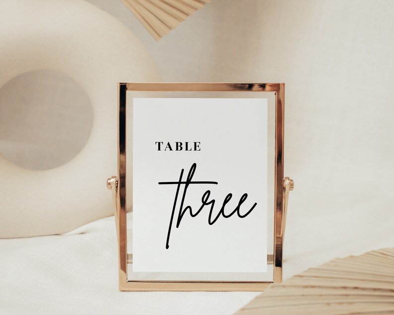 May include: Gold-framed table number sign with a white background and black text that reads "TABLE three".