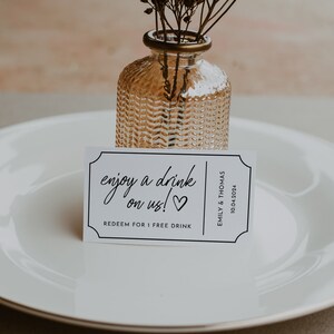 May include: White card with black text that reads "enjoy a drink on us!" with a heart symbol. The card is on a white plate with a small glass vase with dried flowers.