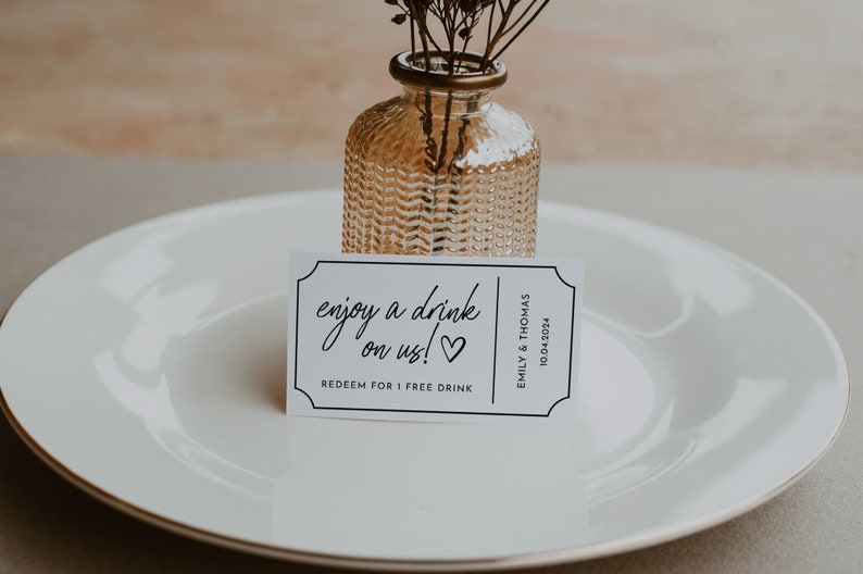 May include: White card with black text that reads "enjoy a drink on us!" with a heart symbol. The card is on a white plate with a small glass vase with dried flowers.
