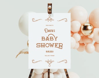 May include: White sign with brown text announcing "Welcome to Dakota's Baby Shower" with the date "10.09.2024" and location "Dallas, Texas". The sign is on a wooden easel with a pink and gold balloon arch behind it.