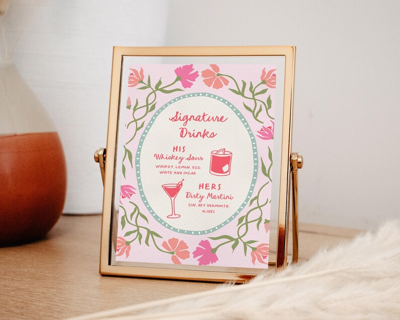 Puede incluir: Un letrero con marco dorado con fondo rosa muestra "Signature Drinks". Enumera "His" Whiskey Sour y "Hers" Dirty Martini, con ilustraciones de c&oacute;cteles. Dise&ntilde;os florales y de vid rosas y verdes rodean el texto.