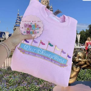 May include: A light pink sweatshirt with the embroidered text "Walt Disney World" and "The Most Magical Place on Earth." The sweatshirt also features a small tag that says "Little Bit of Magic on Main."