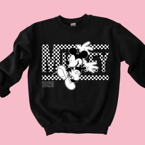 Puede incluir: Sudadera negra con un dise&ntilde;o a cuadros blancos y una imagen en blanco y negro de Mickey Mouse. El texto "Mickey" est&aacute; escrito en letras blancas con un contorno negro. El a&ntilde;o "1928" est&aacute; escrito en blanco debajo de la imagen.