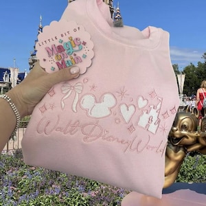 Puede incluir: Sudadera rosa claro con dise&ntilde;os de Disney bordados en blanco, incluyendo orejas de Mickey Mouse, corazones y un castillo. El texto "Walt Disney World" est&aacute; bordado debajo. Una etiqueta con el texto "Little Bit of Magic on Main" est&aacute; adjunta.
