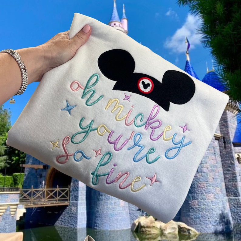 K&ouml;nnte beinhalten: Wei&szlig;er Pullover mit einem regenbogenfarbenen Stickmuster, auf dem "Oh Mickey, you're so fine" steht, und einem schwarzen Mickey-Maus-Kopf mit einem roten Kreis.