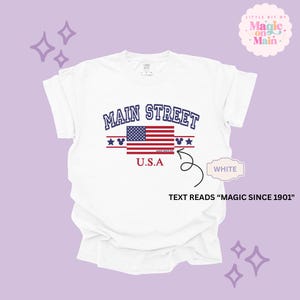 May include: A white short-sleeved t-shirt featuring the text MAIN STREET in blue, a graphic of the American flag, and the text U.S.A in red. Below the flag, small text reads MAGIC SINCE 1901. The design includes blue stars and mouse ear icons.