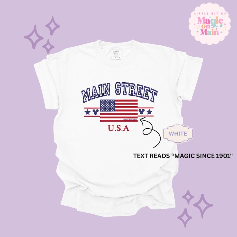 May include: A white short-sleeved t-shirt featuring the text MAIN STREET in blue, a graphic of the American flag, and the text U.S.A in red. Below the flag, small text reads MAGIC SINCE 1901. The design includes blue stars and mouse ear icons.