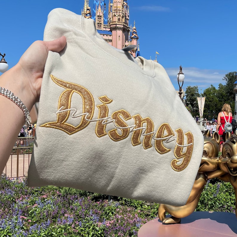 K&ouml;nnte beinhalten: Ein wei&szlig;er Pullover mit einem goldenen Glitzer-&bdquo;Disney&ldquo;-Logo, das auf der Vorderseite gestickt ist. Der Pullover ist gefaltet und vor einem Schloss gehalten.